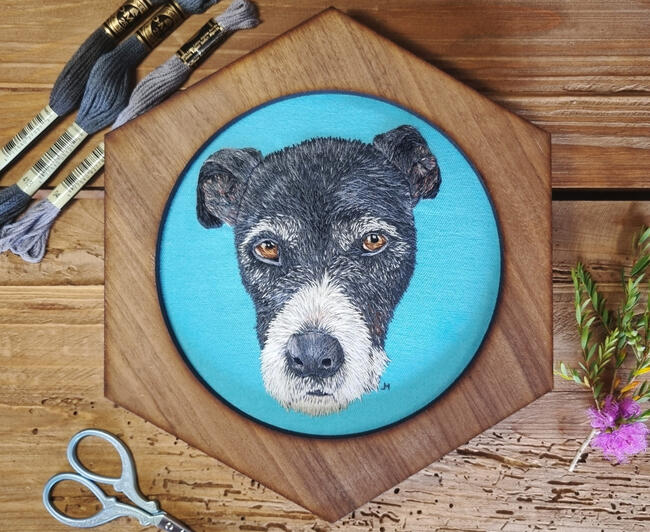 Hand embroidered portrait of black and white dog with sad, brown eyes on light blue fabric in a hexagonal frame