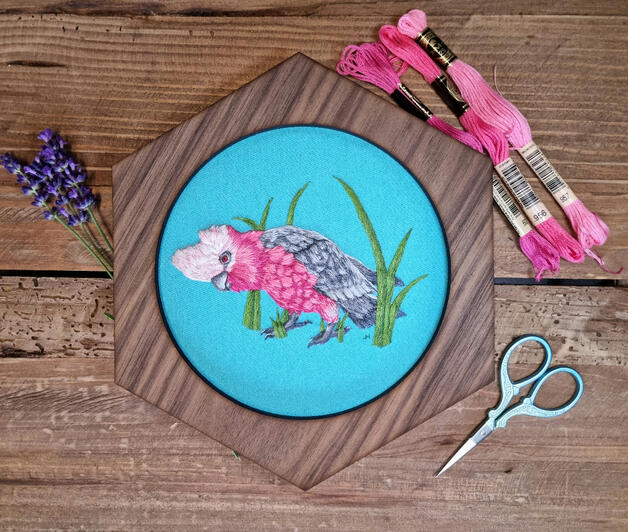 Hand embroidered portrait of a galah standing in grass on light blue fabric in a hexagonal frame