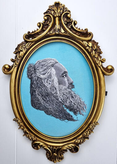 Hand embroidered greyscale portrait of Taika Waititi in an ornate gold frame on light blue fabric
