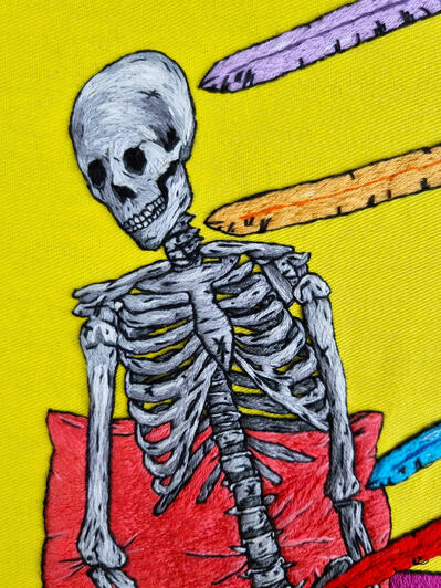 A hand embroidered art piece with a grayscale skeleton on yellow fabric - an orange sword is pointed at its neck and there is a pink pillow behind it.