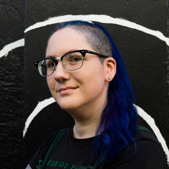 A woman with glasses, shaved sides of head and long blue, purple wavy hair. Her septum is pierced and she is smiling slightly at the camera